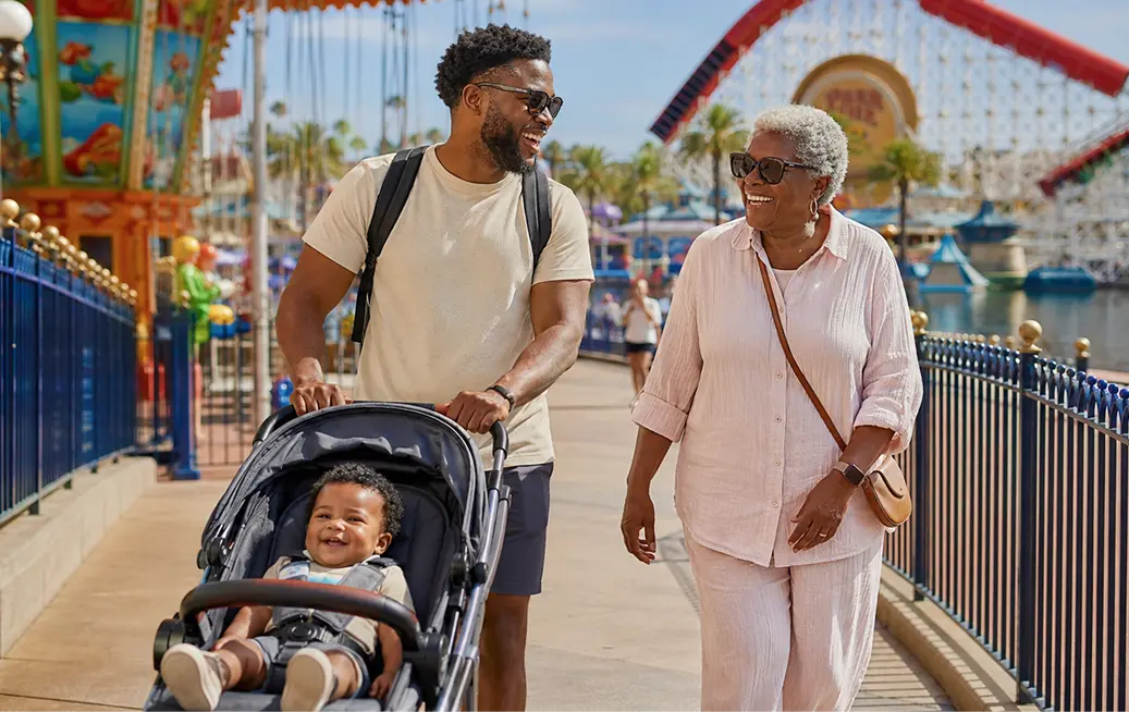Father pushing a smiling baby in a Cloud of Goods rental stroller along the Paradise Pier boardwalk at Disney California Adventure in Anaheim, walking alongside grandmother, with roller coaster visible in the background.