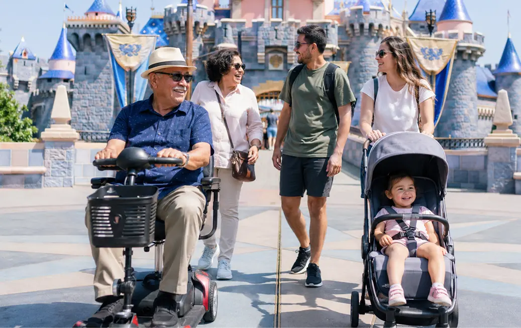 Multigenerational family at Disneyland — grandfather riding a red mobility scooter and young couple pushing a toddler in a stroller, both rentals delivered by Cloud of Goods in Anaheim, with Sleeping Beauty Castle in the background.