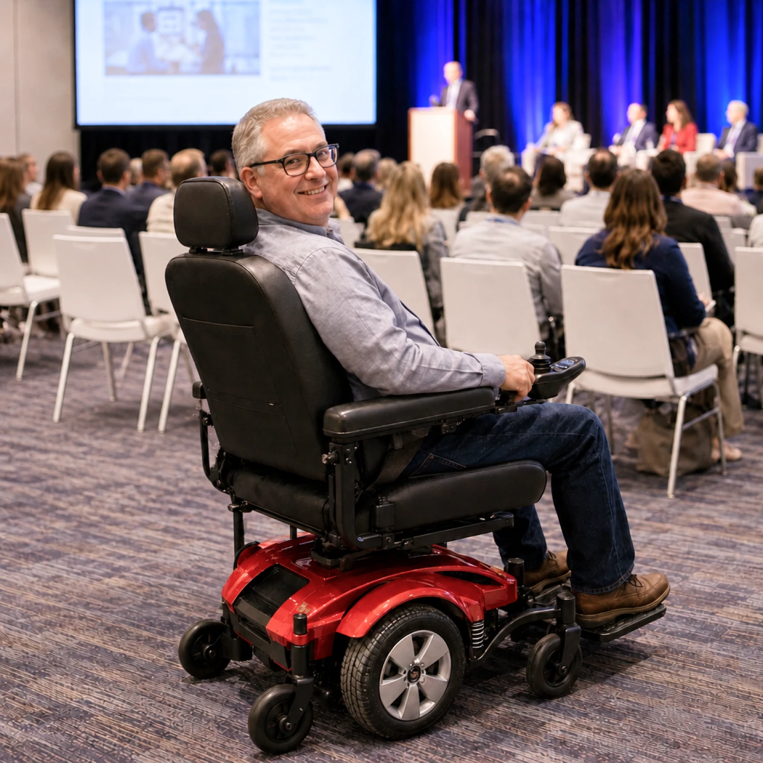 A convention attendee uses a red standard power chair while watching a keynote session in a conference room with another attendee nearby.