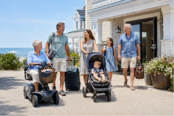 A multigenerational family with a mobility scooter and a stroller arriving at a coastal vacation resort on a sunny day.